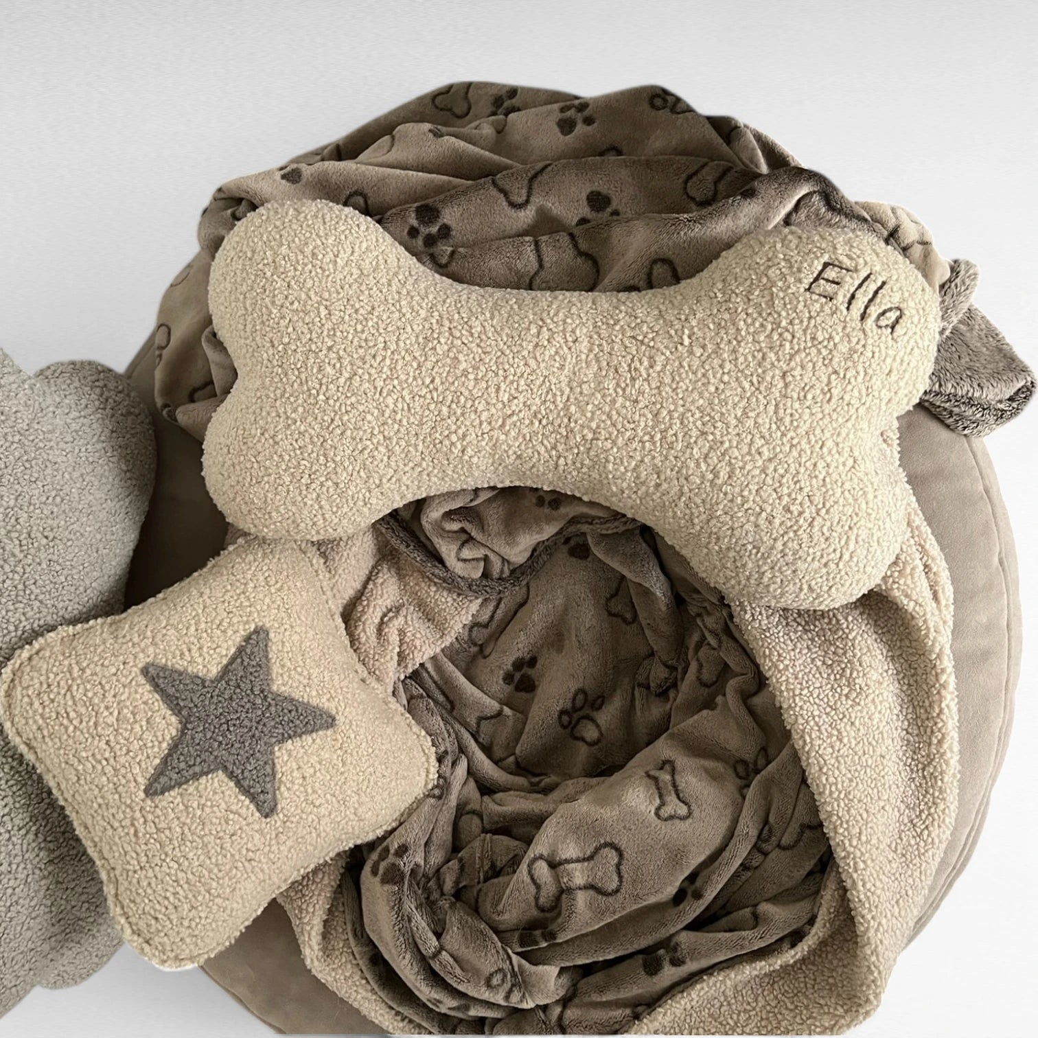 Kuscheliges Hundekissen in Knochenform aus Bouclestoff in beige mit brauner Namensstickerei Ella auf Hundeschlafsack mit Knochenmuster liegend