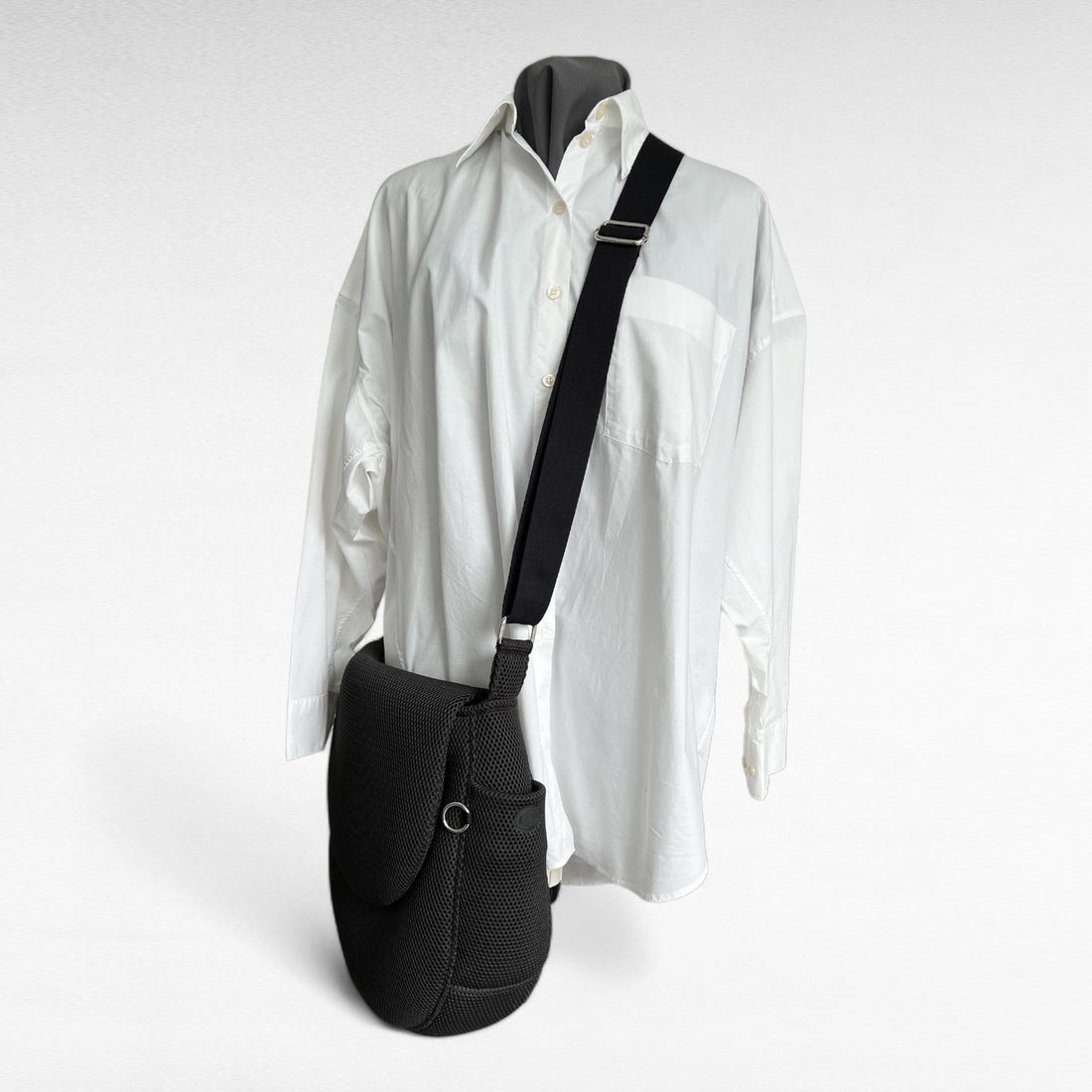 Runde oversized -Tasche mit Taschenklappe aus schwarzem Mesh Material mit schwarzem Gurt und Metallelemente in silber auf Schneiderpuppe mit weißem Hemd