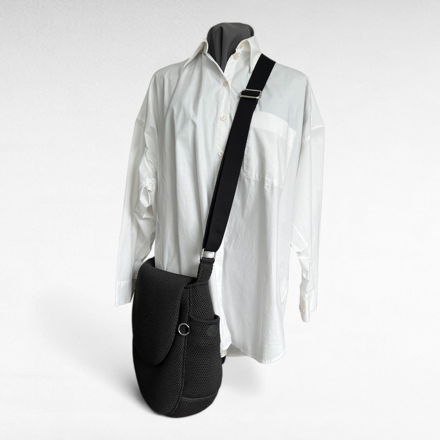 Runde oversized -Tasche mit Taschenklappe aus schwarzem Mesh Material mit schwarzem Gurt und Metallelemente in silber auf Schneiderpuppe mit weißem Hemd