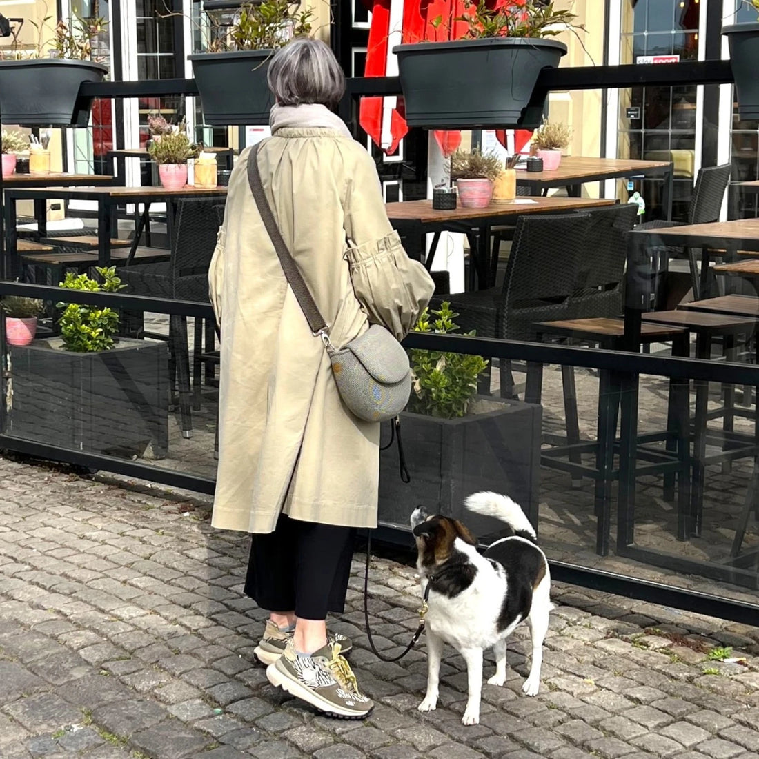 Frau mit extraleichter halbrunder Hundetasche aus beigem Meshmaterial beim Gassigang mit kleinem schwarz weißen Hund an der Leine vor einem Outdoorcafe