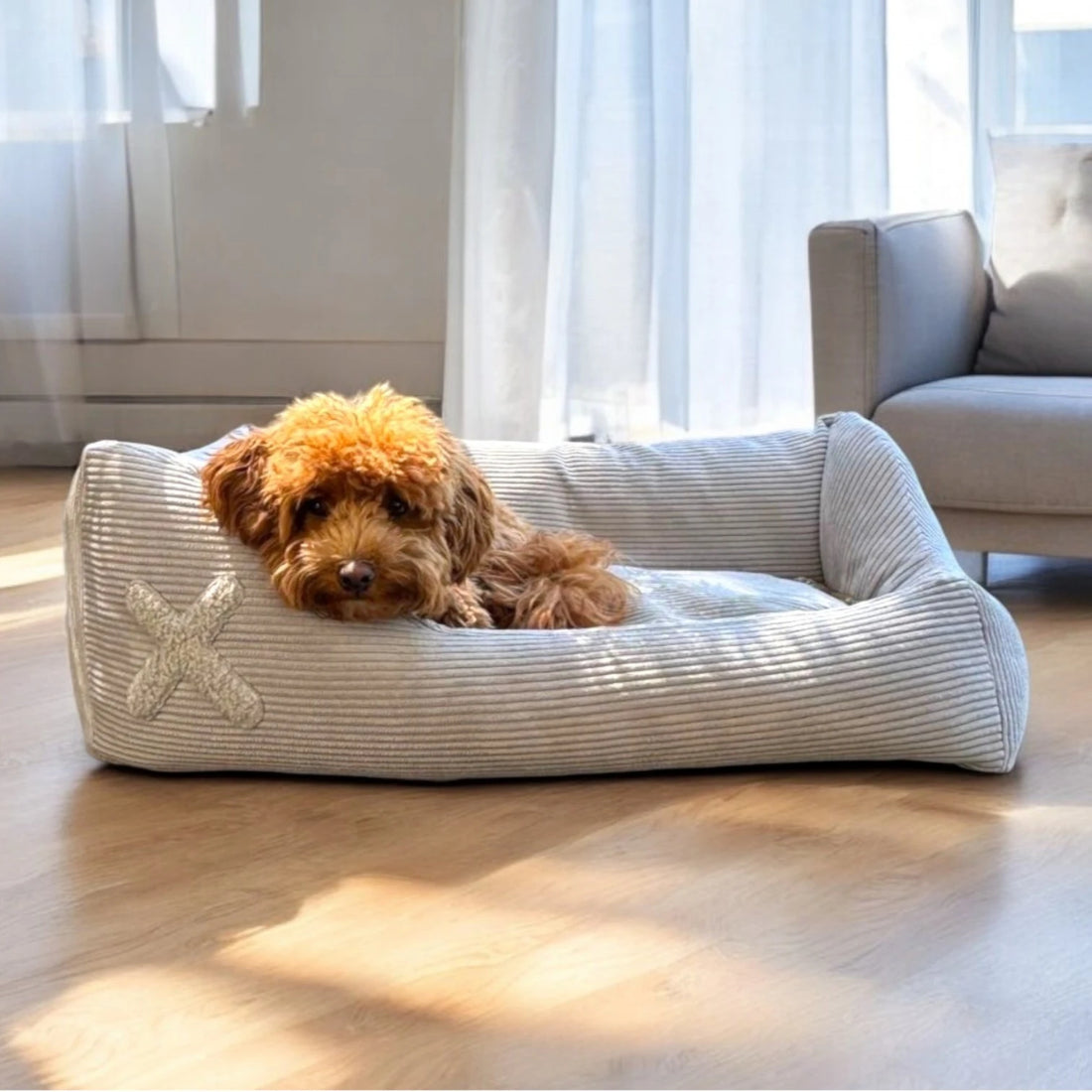 Rechteckiges Hundebett mit asymmetrischen Seitenpolstern und hellgrauem Cordbezug und Plüschapplikation in Kreuzform mit Hund darin liegend in hellem Wohnzimmer