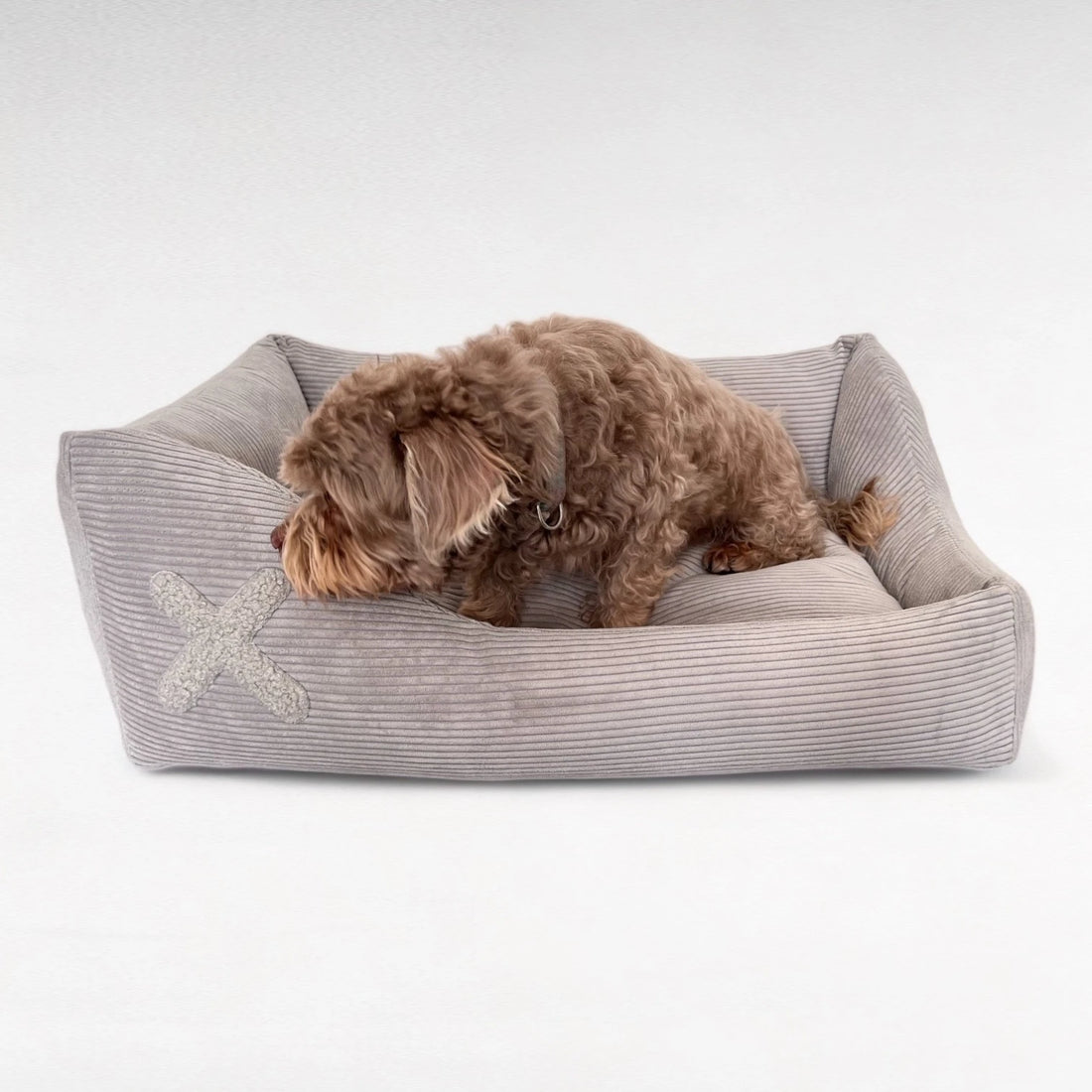 Rechteckiges Hundebett mit asymmetrischen Seitenpolstern und hellgrauem Cordbezug und Plüschapplikation in Kreuzform mit Hund darin sitzend