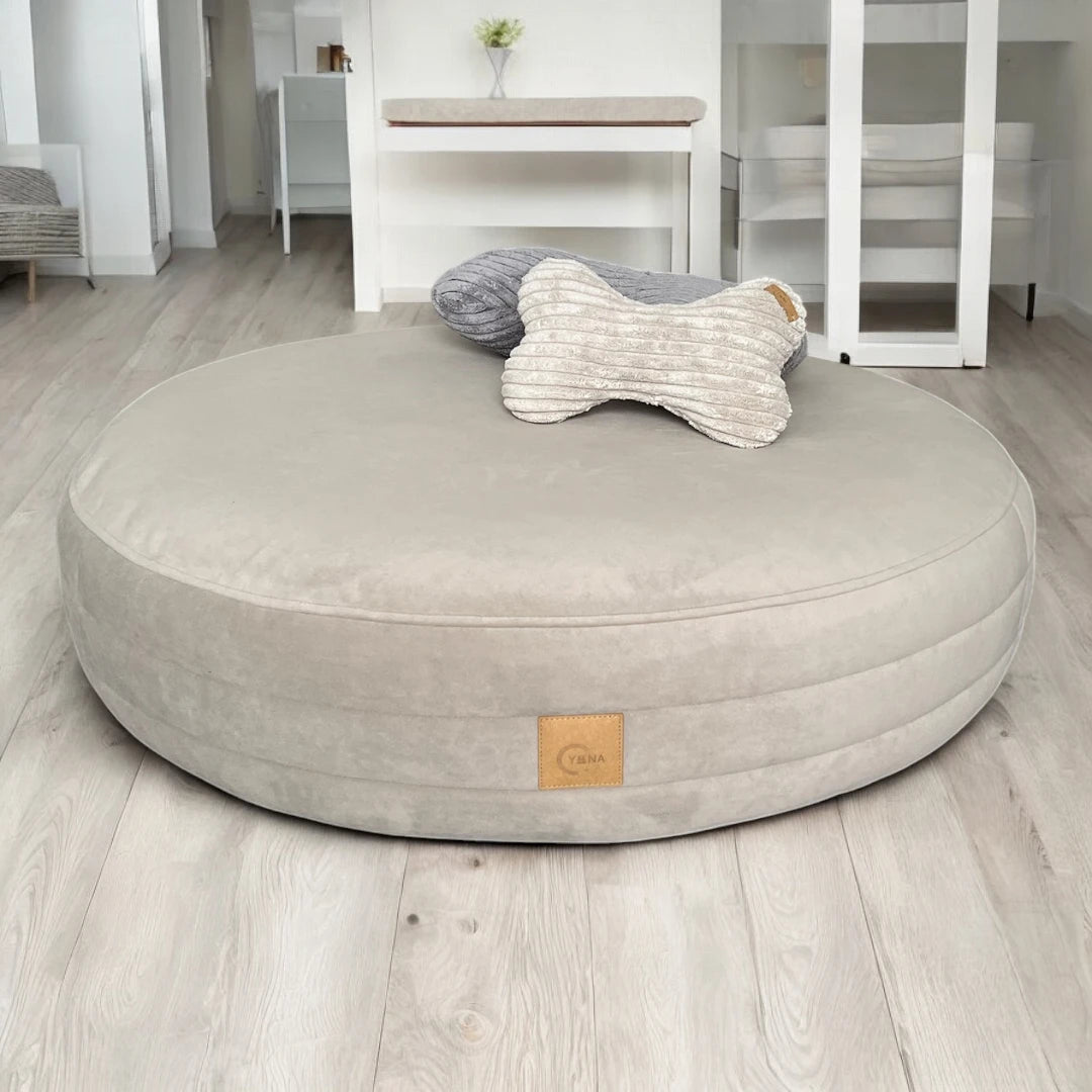 Rundes Hundebett Yumi Farbe greige mit rundherum verlaufenden Steppungen mit Yuna Etikett, sowie 2 kleinen Breitcordkissen auf dem Bett liegend mit Wohnzimmer im Hintergrund