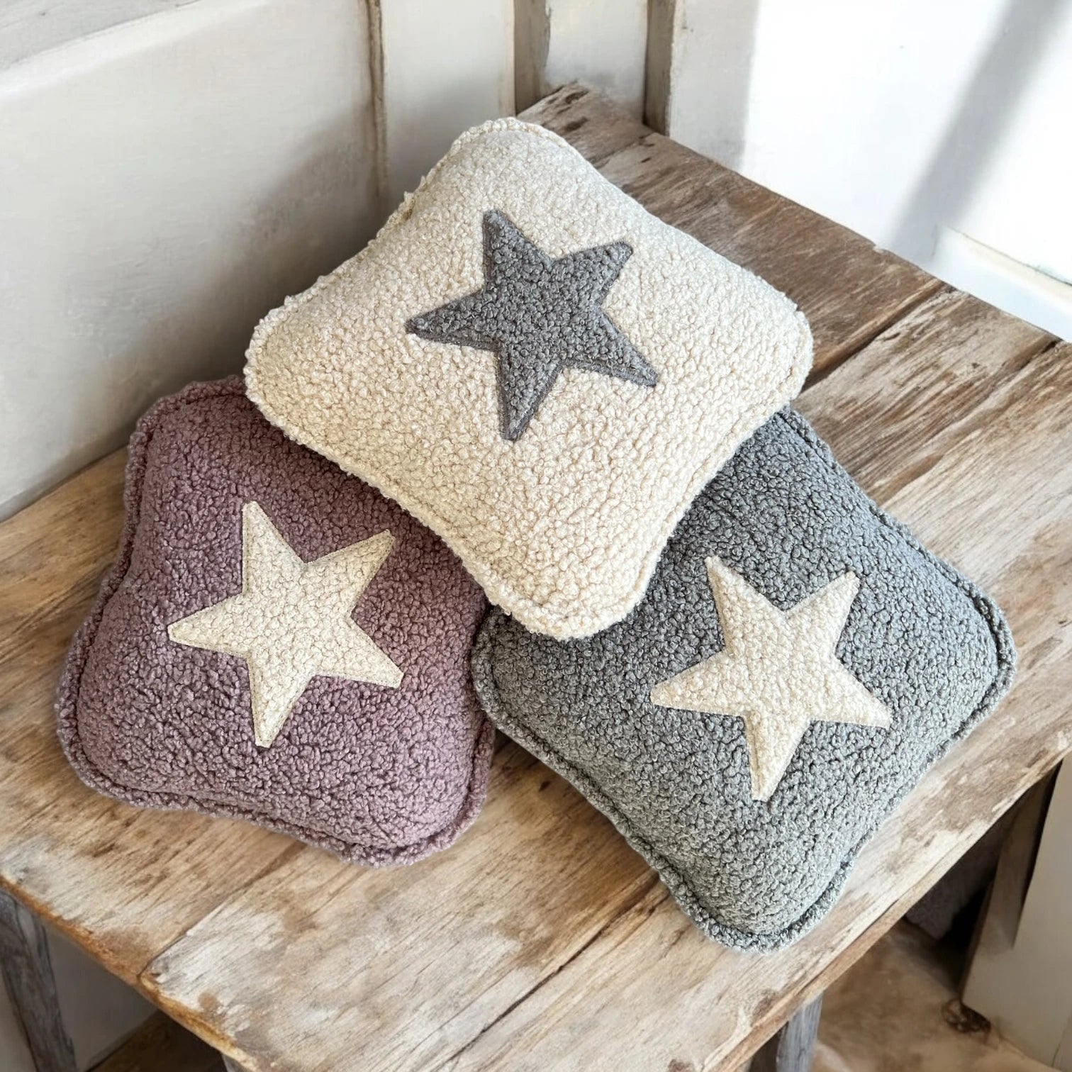 3 quadratische Kissen aus Boucle in beige, altrosa und grau jeweils mit Stern Applikation auf Holztisch