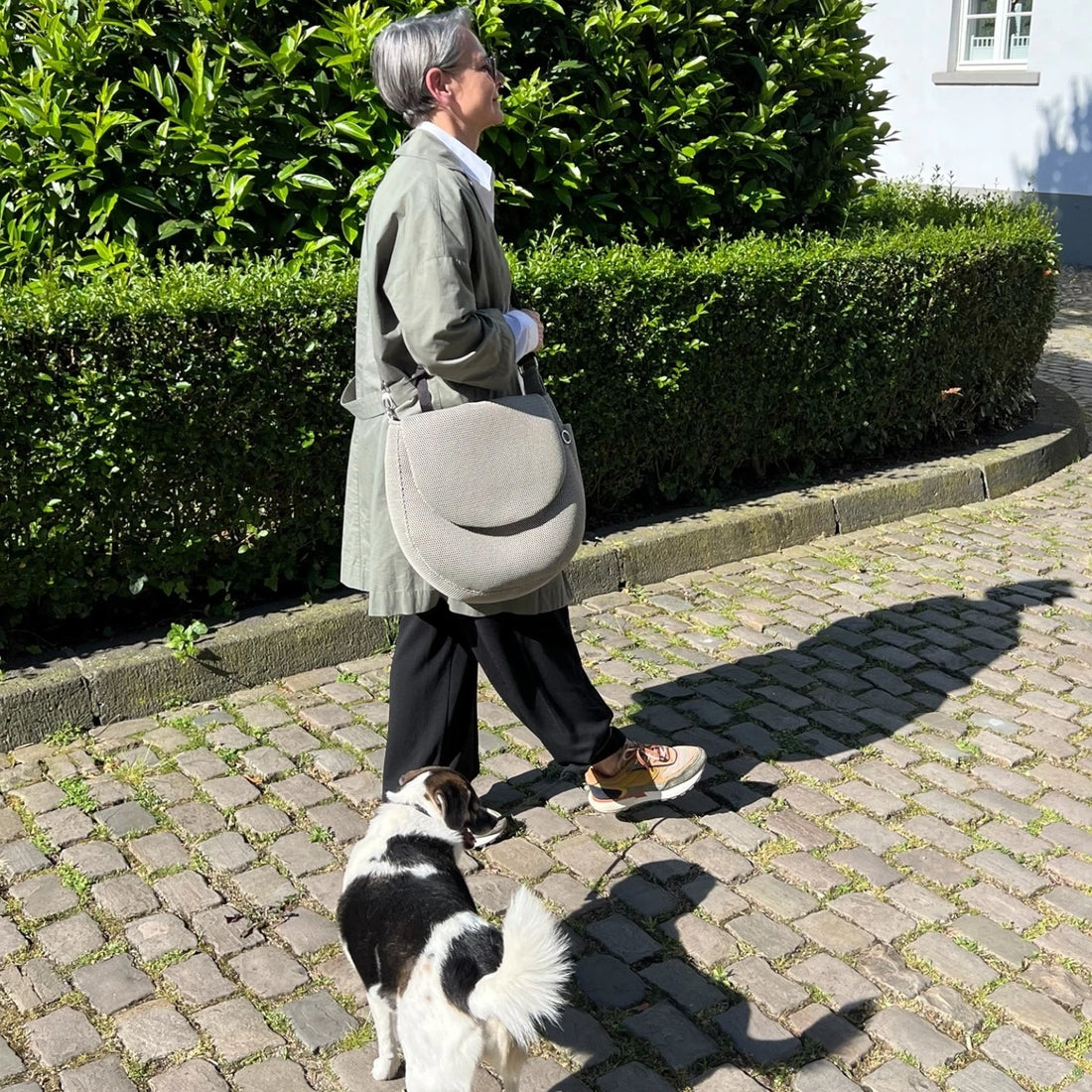 Frau mit Hund beim Spazieren gehen, dabei trägt die Frau eine runde oversized Tasche aus Mesh Material in Farbe beige, im Hintergrund sind grüne Büsche