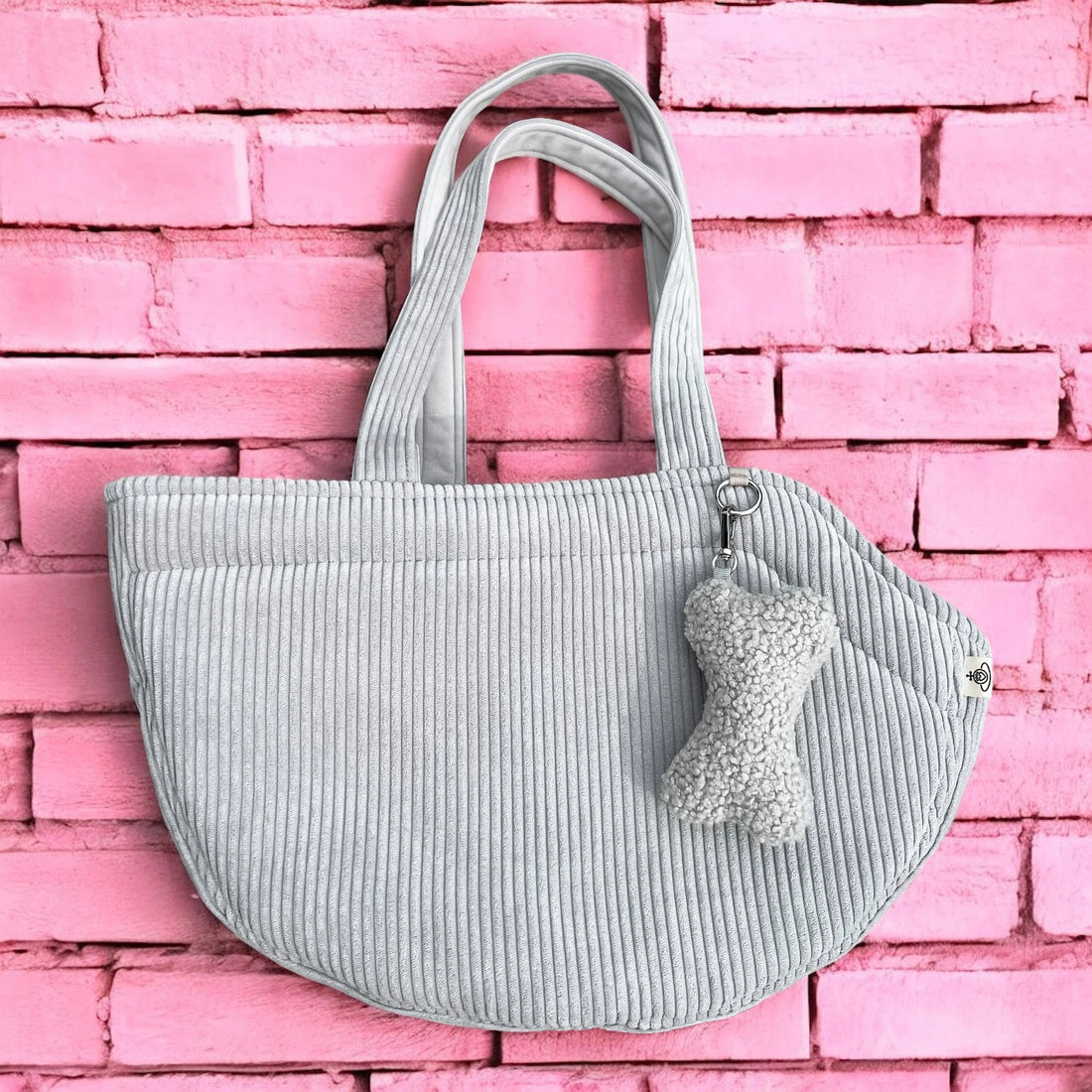 Halbrunde Hundetasche aus Cord hellgrau mit 2 Tragegurten aus Cordstoff, sowie silberner O-Ring und Taschenanhänger in Knochenform in Boucle hellgrau mit pinkfarbenen Hintergrund