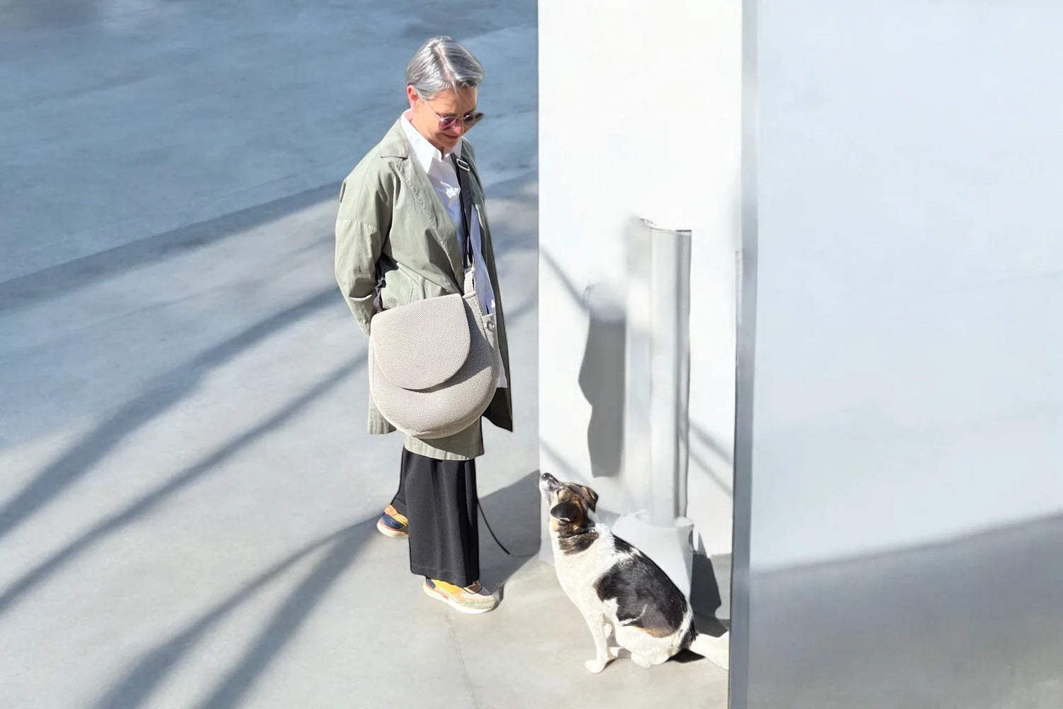 Frau mit großer Crossbody Tasche in der Sonne stehend und ein kleiner Hund sitzt vor ihr heraufschauend