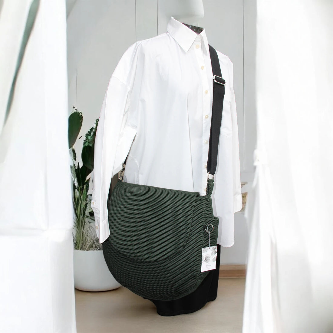 Runde dunkelgrüne oversized Tasche mit Taschenklappe aus Mesh Material mit schwarzem Gurt und Metallelemente in silber auf Puppe mit weißen Hemd mit minimalistischem Hintergrund