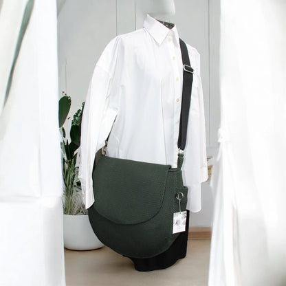 Runde dunkelgrüne oversized Tasche mit Taschenklappe aus Mesh Material mit schwarzem Gurt und Metallelemente in silber auf Puppe mit weißen Hemd mit minimalistischem Hintergrund
